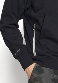 Felpa nera con tasca con zip e logo "DRI-FIT" sul polsino. Presenta polsini a costine, tasca a marsupio e una texture del tessuto liscia.