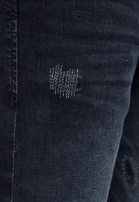 Mörka denimjeans med en liten sydd lapp som reparerar en rivning vid lårområdet.