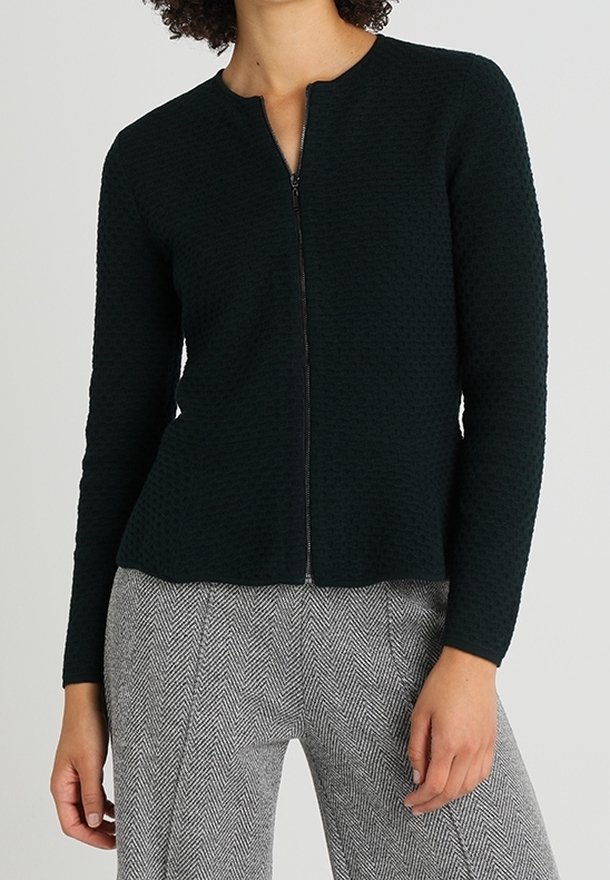 Ciemnozielony sweter rozpinany na zamek z teksturowanym wzorem, okrągłym dekoltem i długimi rękawami, zestawiony z spodniami w kratę hounds-tooth.