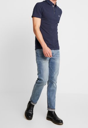 Mężczyzna ubrany w ciemnogranatową koszulkę polo, jasnoniebieskie podwinięte dżinsy i czarne skórzane botki, stojący na tle jednolitego szarego tła.