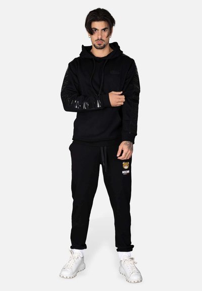 Felpa nera con accenti lucidi sulle maniche, abbinata a pantaloni jogger neri con un logo di un orso. Entrambi sono realizzati in tessuto morbido, modello con sneakers bianche.