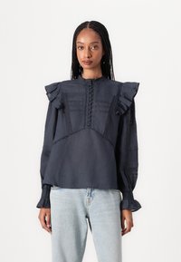 Dark navy blouse with ruffle sleeves, textured detailing, button closure, and embroidered accents. Paired with light blue jeans.