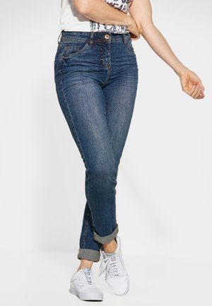 Vrouw draagt opgerolde blauwe spijkerbroek en witte sneakers, staat met één been gekruist over het andere tegen een effen witte achtergrond.
