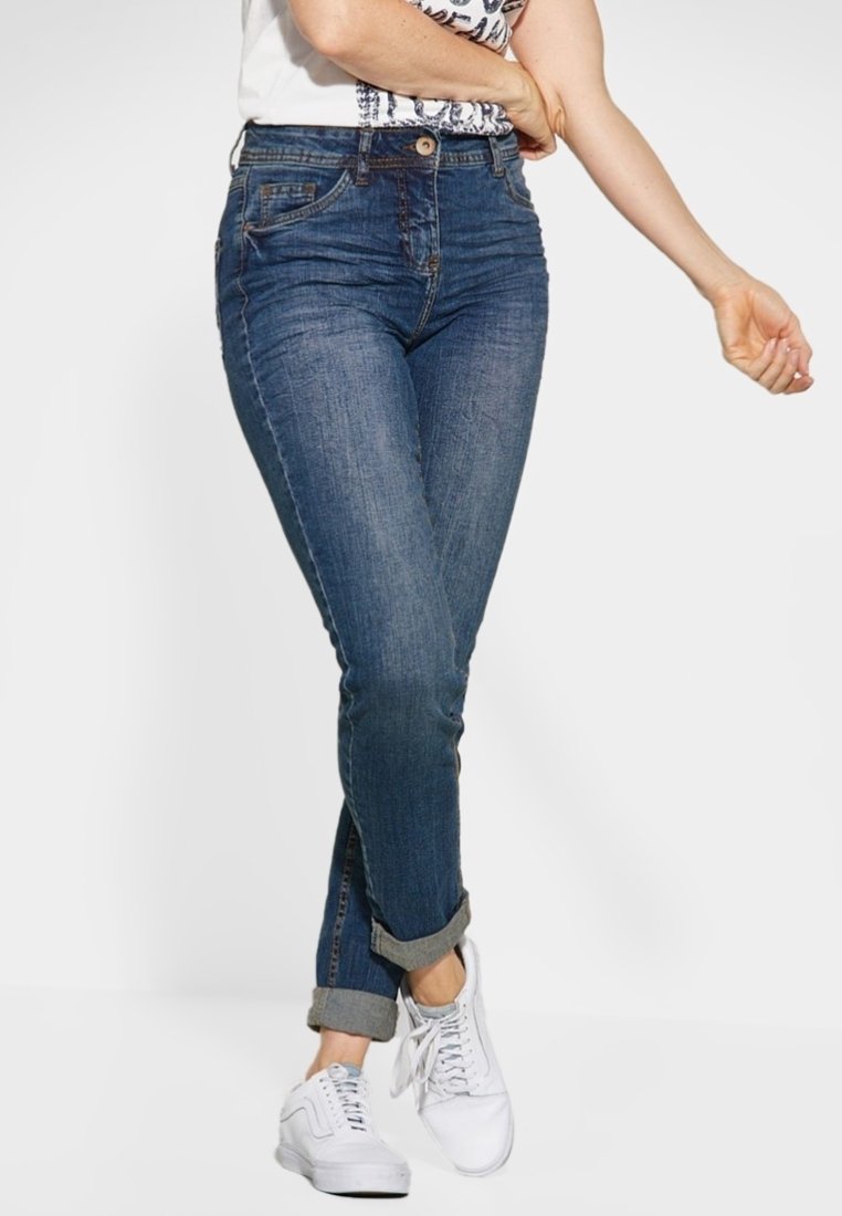 Femme portant un jean bleu retroussé et des baskets blanches, debout avec une jambe croisée sur l'autre sur un fond blanc uni.
