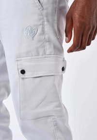 Primo piano di pantaloni cargo grigi chiaro con tasca a toppa e dettagli di bottoni neri, accanto a una mano di pelle scura appoggiata sulla coscia.
