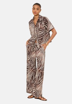 Femme portant une chemise ample à manches courtes imprimée zèbre et un pantalon large assorti avec des sandales noires, debout les mains dans les poches.