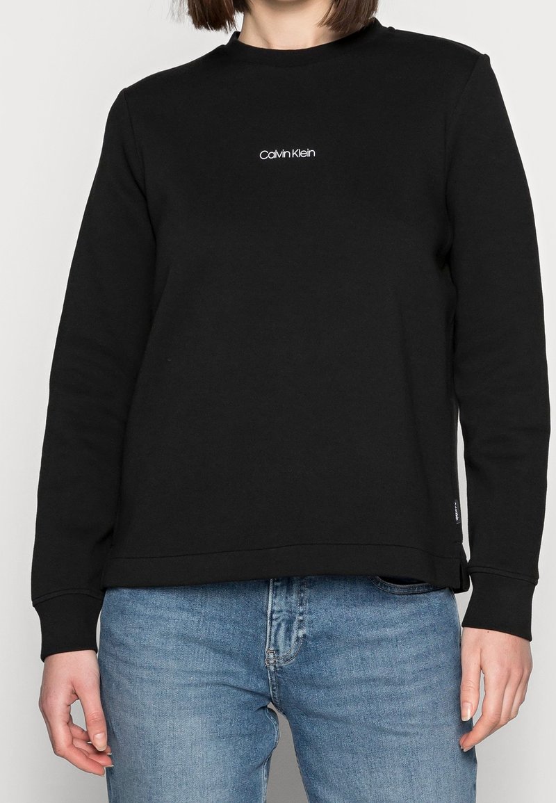 Musta puuvillainen svetari pyöreällä pääntiellä, resorireunuksilla hihoissa ja helmassa. Rinnassa valkoinen "Calvin Klein" -logo.