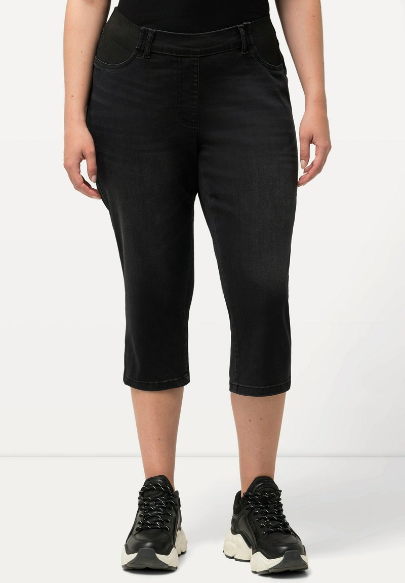 Pantalones capri negros de mezclilla, con una textura suave, bolsillos traseros curvados y una cintura ajustada. Llevados con zapatillas negras.