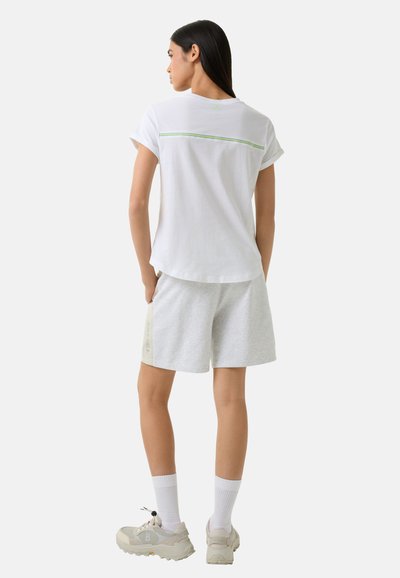 Hvid kortærmet skjorte med grøn detalje, parret med grå shorts med sidebranding. Modellen har lyse sko og hvide sokker på.