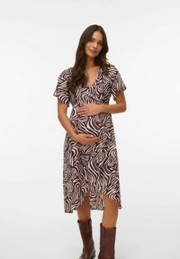 Vestido de maternidad con escote en pico profundo, mangas cortas y un patrón ondulado en blanco y púrpura. Tiene un dobladillo asimétrico y se combina con botas marrones.