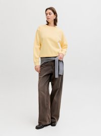 Femme debout portant un pantalon ample marron, un haut à manches longues jaune, un pull gris noué à la taille, et des chaussures noires, sur un fond uni.