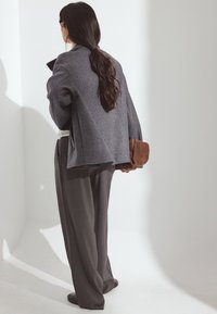 Pelēks pārāk liels žakets ar teksturētu audumu, saskaņots ar plašām tumši pelēkām biksēm. Brūns plecu somiņš turēts rokā. Minimālisma dizains.