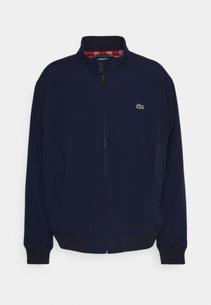 Laivasilkkinen takki, joka on valmistettu pehmeästä kankaasta, jossa on korkea kaulus, vetoketjukiinnitys, sivutaskut ja ruudullinen vuori. Rinnassa on vihreä logo.