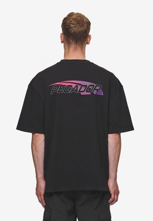 Uomo che indossa una t-shirt nera oversize con la scritta "PEGADOR" in testo stilizzato rosa e viola sulla parte alta della schiena, in piedi davanti a uno sfondo bianco.