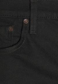 Lähetysteksti mustasta denimgofriikista lähikuvassa, jossa on etutasku, ruskeat metallin niitit ja kontrastoiva ompelu. Sujuva rakenne, jossa on hieno kiilto.