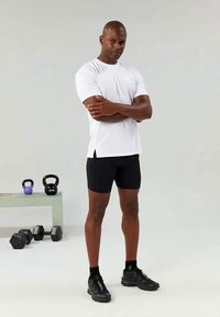 Tricou athletic alb cu fante laterale, purtat cu pantaloni şort negri. Bărbat în picioare cu brațele încrucișate, înconjurat de kettlebell-uri și gantere.