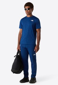 Muž v modrém tričku a kalhotách North Face, s černými slunečními brýlemi, držící černý batoh, stojící na jednobarevném bílém pozadí.