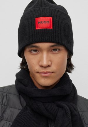 Zwarte geribbelde gebreide beanie met een rood rechthoekig etiket met de tekst "HUGO"; gedragen met een bijpassende zwarte gebreide sjaal.