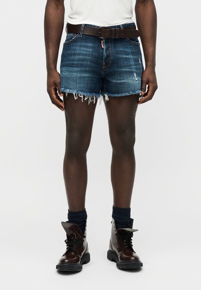 Άνδρας με σκουρόχρωμα φθαρμένα σορτς από τζιν, καφέ ζώνη, σκουρόχρωμες κάλτσες και καφέ μποτάκια με κορδόνια, μπροστά σε απλό φωτεινό φόντο.