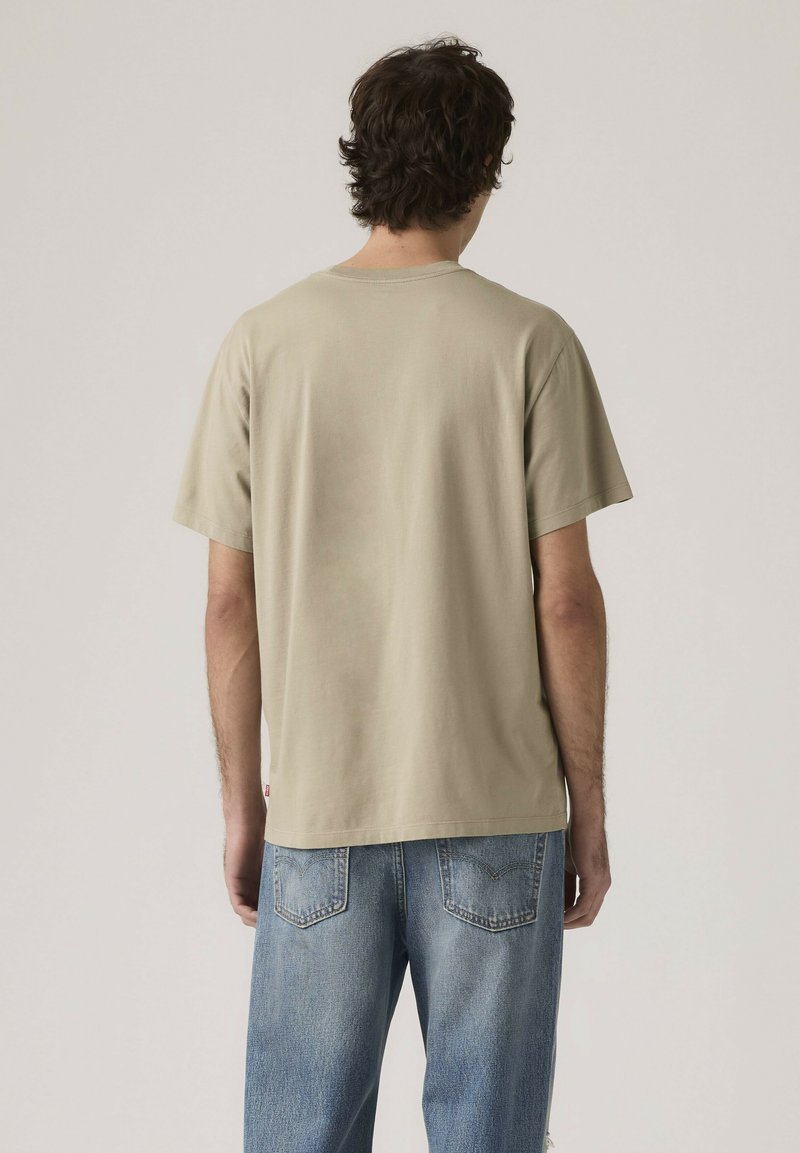 Uomo visto da dietro che indossa una t-shirt beige a maniche corte e jeans blu, su sfondo chiaro uniforme.