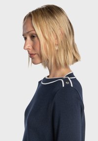 Pull bleu marine en tissu texturé, avec un col blanc bordé d'un liseré et deux boutons décoratifs sur l'épaule.