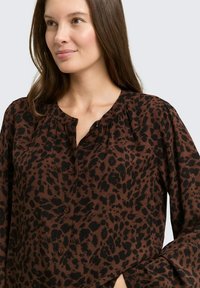 Blouse marron avec un imprimé animalier noir, boutons sur le devant, détails froncés au niveau de l'encolure, et manches longues à coupe décontractée.