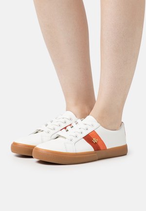 Białe niskie trampki z brązowymi gumowymi podeszwami, pomarańczowo-czerwonymi bocznymi paskami oraz złotym logo, noszone na bose stopy na białym tle.