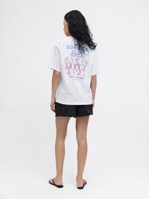 Kvinde med langt krøllet hår står med ryggen til iført en hvid T-shirt med tre hummere og tekst, sorte shorts og sandaler på hvid baggrund.
