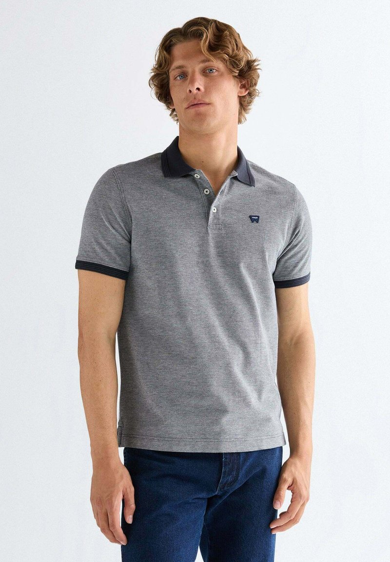 Ung mand med bølget hår iført en grå poloshirt med mørk krave og ærmekanter, kombineret med mørkeblå jeans, stående mod en hvid baggrund.