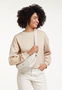 Sweat-shirt beige à manches larges, associé à un sac bandoulière clair arborant un branding minimal. Le mannequin se tient devant un fond neutre.