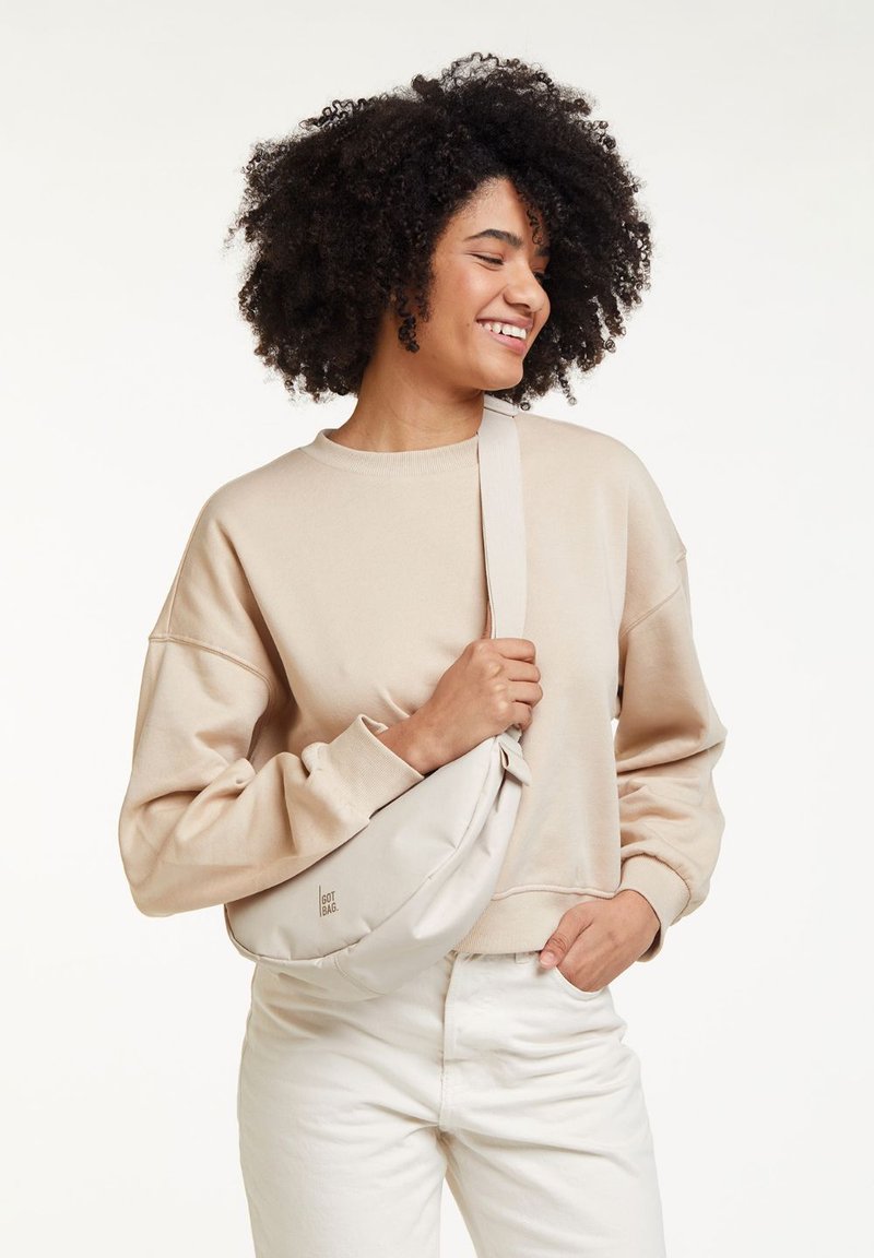 Sweat-shirt beige à manches larges, associé à un sac bandoulière clair arborant un branding minimal. Le mannequin se tient devant un fond neutre.