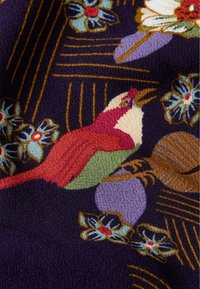 Textil med en mörk lila bakgrund och färgglada blommor och fågelmönster, inklusive en röd och grön fågel och blå blommor.