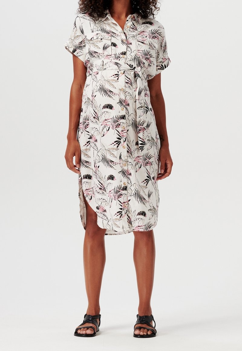 Femme portant une robe courte aux genoux, à manches courtes, boutonnée sur le devant, avec une ceinture à nouer et un imprimé botanique, associée à des sandales noires.