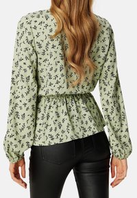 Blouse à motif floral vert avec des manches longues, taille froncée et poignets élastiques. Assortie à un pantalon noir ajusté.
