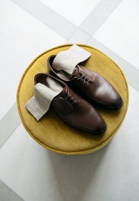 Hnědé kožené boty na tkaničky, umístěné na kulatém, žlutě čalouněném taburetu. Světle béžové ponožky s modrými puntíky leží přes boty.