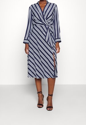 Femme portant une robe portefeuille rayée bleu marine et blanche à manches longues avec une fente latérale, associée à des sandales à talons hauts noires à bride cheville.