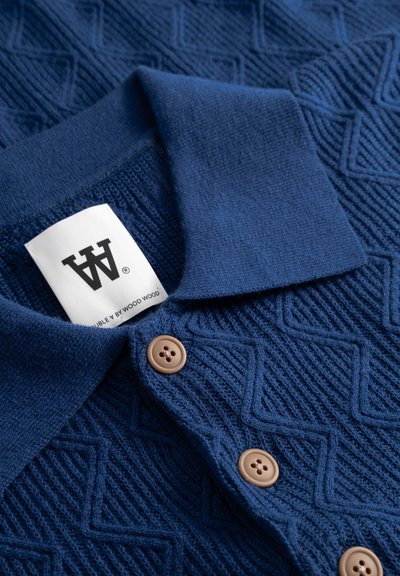 Polo en maille bleue présentant un motif texturé en losanges, avec un col et des boutons en plastique marron, portant une étiquette indiquant la marque.