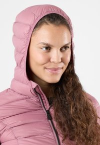 Lys pink puffjakke med hætte, der har et quiltet mønster, lynlås og blød tekstur. Båret af en person med langt, krøllet hår.