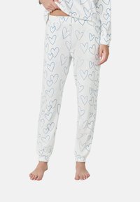 Persoon in witte pyjamabroek met een patroon van met blauwe omlijnde harten, staande op blote voeten tegen een effen achtergrond.