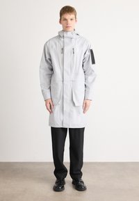 Căciulă ușoară gri cu glugă, două buzunare frontale și o pată de umăr neagră, având un design drept și material impermeabil. Pantaloni și pantofi negri.
