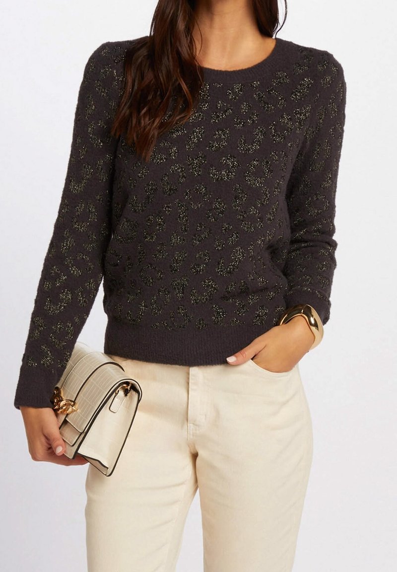 Femme portant un pull gris foncé à imprimé léopard, un pantalon beige, tenant un sac à main crème, et portant un bracelet doré au poignet.