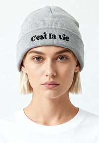 Jeune personne aux cheveux blonds courts portant un bonnet gris en tricot brodé de « c'est la vie » et un t-shirt blanc uni, fond neutre.