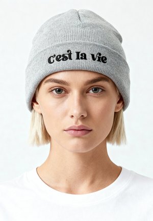 Young person with short blonde hair wearing a gray knit beanie embroidered with "c'est la vie" and a plain white shirt, neutral background.