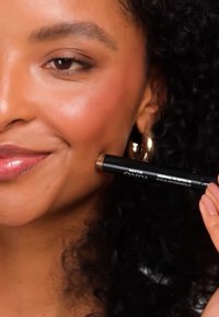 Zwarte make-up stick dicht bij de wang gehouden. Het heeft een draaibaar ontwerp met een afgeronde punt. Het model heeft een warme huidtint en glanzende lippen.