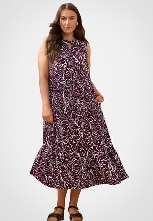 Kobieta ubrana w fioletową sukienkę bez rękawów sięgającą kostek z białym abstrakcyjnym wzorem oraz czarne sandały na kostkę ze paskiem, stojąca z jedną ręką w kieszeni.