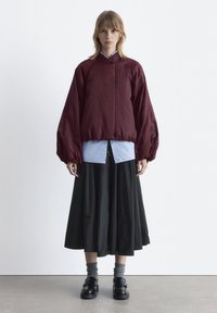 Burgunderfarbener Puffer-Jacke mit übergroßen Ärmeln, darüber ein hellblaues Hemd, kombiniert mit einem schwarzen Rock und glänzenden schwarzen Schuhen.