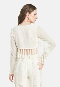 Weißes, gestricktes Crop-Top mit langen Ärmeln, einem Fransenabschluss und einem offenen Rücken. Die Textur ist leicht und luftig, mit einem lockeren Schnitt.