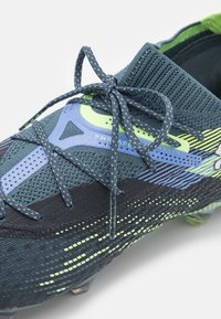 Scarpa da calcio di colore blu scuro, con una parte superiore texturizzata, dettagli in verde neon e lacci grigi. Presenta strisce diagonali e una forma aderente.