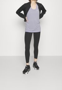 Kvinna står med händerna på höften, klädd i en svart jacka, lavendelfärgad linne, svarta leggings och svarta sportskor.