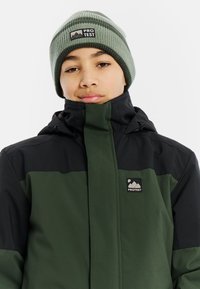 Grüne und schwarze Jacke mit hohem Kragen und Reißverschluss, ausgestattet mit einem Logo-Patch. Strickmütze in Grün mit Streifen. Glatte Textur.